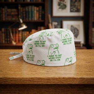 May include: White surgical scrub cap with a repeating pattern of green cartoon frogs and the text "I HAVE TO SAY WEIRD STUFF OR I'LL DIE." The cap has ties for a secure fit and is made of a soft, lightweight fabric.