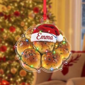 May include: A clear acrylic hanging ornament shaped like a cluster of golden-brown dinner rolls. A red Santa hat with white trim sits on top of the rolls, featuring the name Emma in red script. A string of colorful fairy lights wraps around the rolls.