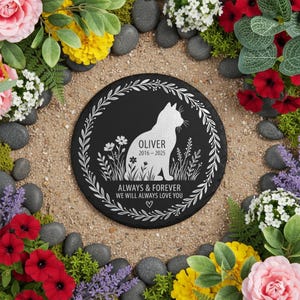 May include: A black, round memorial stone with a white cat silhouette, surrounded by a floral wreath. The text reads "OLIVER 2016-2025" and "ALWAYS & FOREVER WE WILL ALWAYS LOVE YOU". The stone is surrounded by flowers and pebbles.