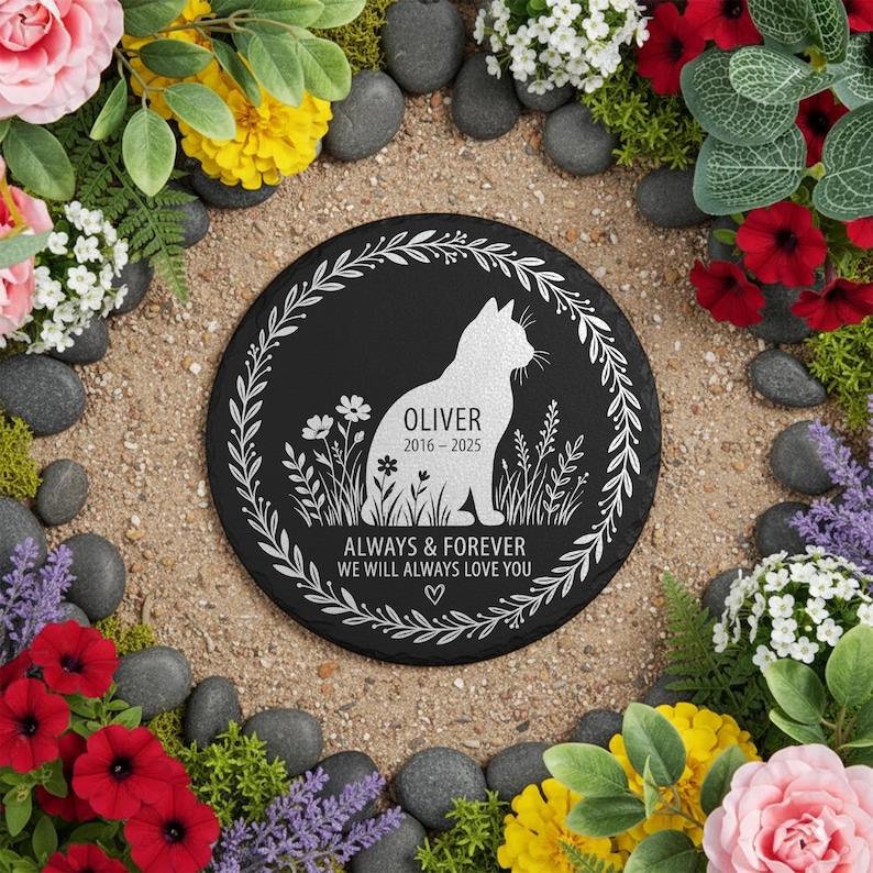 May include: A black, round memorial stone with a white cat silhouette, surrounded by a floral wreath. The text reads "OLIVER 2016-2025" and "ALWAYS & FOREVER WE WILL ALWAYS LOVE YOU". The stone is surrounded by flowers and pebbles.