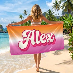 May include: A beach towel with the name "Alex" in a retro font, displayed against a gradient of orange, pink, and white. The towel is held by a person on a sandy beach with palm trees and blue water in the background.