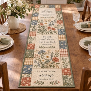 May include: A rectangular fabric table runner featuring a patchwork border of floral and checkered squares in muted tones. The center panel displays three sections with botanical illustrations and the text: I can do all things through Christ who strengthens me, Philippians 4:13; Be still, and know that I am God, Psalm 46:10; and I AM WITH YOU always, Matthew 28:20.