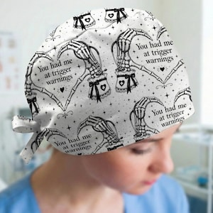 May include: A white scrub cap featuring a repeating pattern of skeleton hands forming heart shapes, black bows, small stars, and the text You had me at trigger warnings. The design includes medical-themed accents and a tie-back closure.
