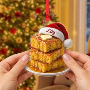 Op de afbeelding: Een doorzichtige acryl kerstornament in de vorm van een stapel van drie goudbruine maïsbroodvierkantjes op een wit bordje. Het bovenste stukje heeft een klontje boter en een rode kerstmuts met de naam Lily in rode letters op de witte rand.