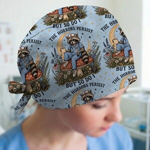 May include: A light blue scrub cap featuring a repeating pattern of a raccoon dressed in blue medical scrubs and a nurse cap, sitting on a mushroom. The text "BUT SO DO I THE HORRORS PERSIST" is printed around the raccoon, surrounded by flowers.