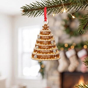 May include: A tiered gingerbread-style Christmas tree ornament hanging from a red ribbon. The ornament features six levels, each decorated with white icing, small pearl-like beads, and seashell accents. The names Anthony, Isabella, Alex, Evelyn, Charlotte, and the year 2026 are printed on the tiers, topped with a small gingerbread person.