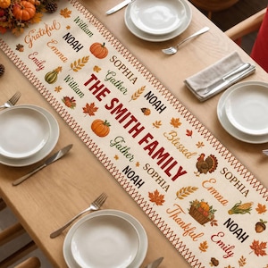 May include: A cream-colored table runner featuring a repeating autumn-themed pattern of pumpkins, fall leaves, wheat stalks, and turkeys. The text includes the phrase THE SMITH FAMILY along with various names like Noah, Emma, and Sophia in different fonts.