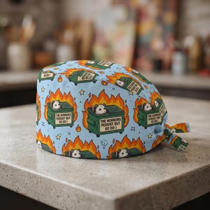 May include: A light blue scrub cap with a repeating pattern of green dumpsters engulfed in flames, each containing a possum. The text "THE HORRORS PERSIST BUT SO DO I" is printed on the dumpsters. The design also includes yellow stars.