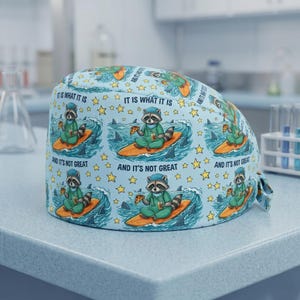 May include: A light blue surgical cap with a repeating pattern of a raccoon surfing on a surfboard, eating pizza, and surrounded by sharks and stars. Text on the cap reads "IT IS WHAT IT IS" and "AND IT'S NOT GREAT."