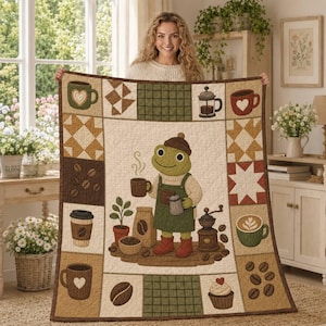 May include: A rectangular quilt featuring a central illustration of a green frog wearing a brown hat, cream sweater, and green apron, holding a coffee mug. The border displays coffee-themed motifs including mugs, beans, and a grinder.