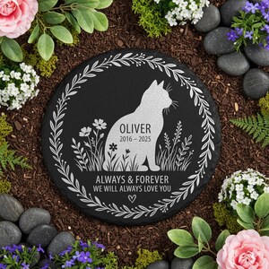 May include: A black, round memorial stone with a white cat silhouette, flowers, and the name "Oliver" with dates. The text "ALWAYS & FOREVER WE WILL ALWAYS LOVE YOU" is also included. The stone is surrounded by a floral wreath.