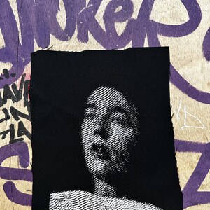Op de afbeelding: Een zwarte vierkante patch met een gedetailleerd wit portret van een menselijk gezicht. De afbeelding is gemaakt met een reeks fijne, parallelle lijnen. De patch is bevestigd op een houten oppervlak met paarse graffiti.