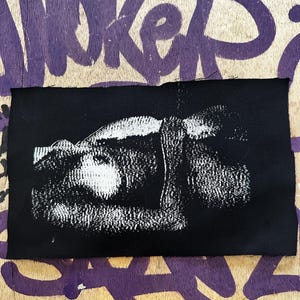 Könnte beinhalten: Ein schwarzer rechteckiger Aufnäher mit einem weißen grafischen Design. Der Aufnäher befindet sich auf einer Holzoberfläche mit lila Graffiti. Die Grafik scheint ein abstraktes Bild zu sein.