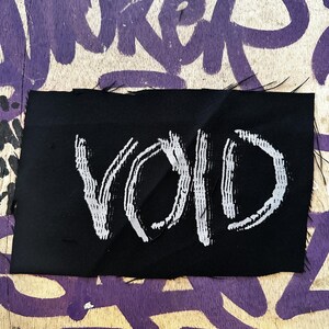 Könnte beinhalten: Ein schwarzer Aufnäher mit dem Wort "VOID" in weißer, pinselartiger Schrift. Der Aufnäher befindet sich vor einem Hintergrund aus verwittertem Holz mit lila Graffiti.