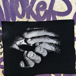Op de afbeelding: Een zwarte stoffen patch met een wit, abstract ontwerp. De patch is rechthoekig met een rafelige rand. De achtergrond is een houten oppervlak met paarse graffiti.