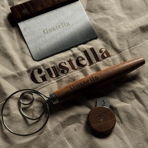 Peut inclure: Un grattoir &agrave; p&acirc;te en acier inoxydable avec un manche en bois fonc&eacute;, un fouet &agrave; p&acirc;te en m&eacute;tal avec un long manche en bois et une petite lame &agrave; pain circulaire en bois avec une lame de rasoir. Chaque objet porte le nom Gustella grav&eacute;.