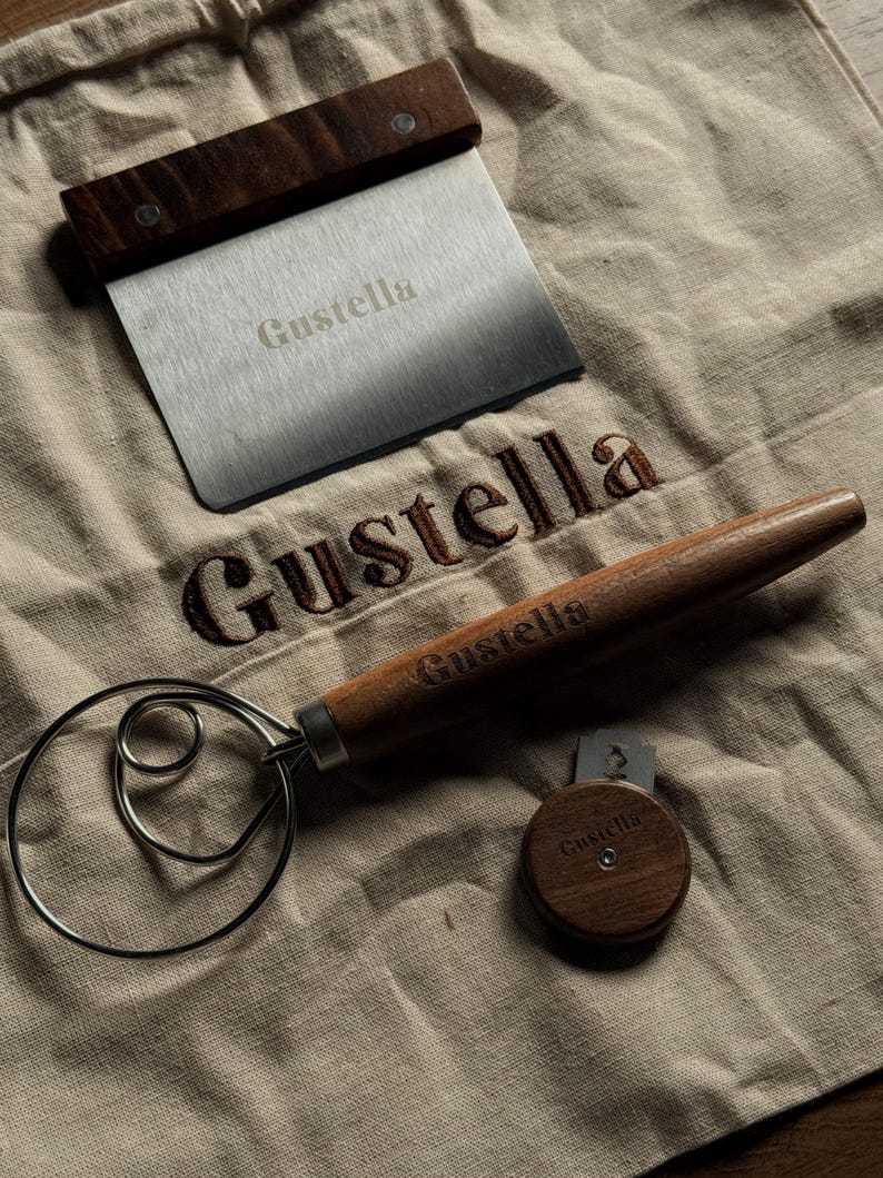 Peut inclure: Un grattoir &agrave; p&acirc;te en acier inoxydable avec un manche en bois fonc&eacute;, un fouet &agrave; p&acirc;te en m&eacute;tal avec un long manche en bois et une petite lame &agrave; pain circulaire en bois avec une lame de rasoir. Chaque objet porte le nom Gustella grav&eacute;.