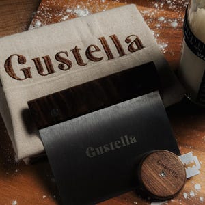 Peut inclure: Un linge en lin beige avec le texte brod&eacute; en marron Gustella, une corne &agrave; p&acirc;tisserie en m&eacute;tal noir avec un manche en bois fonc&eacute; portant le texte grav&eacute; Gustella, et une lame &agrave; pain ronde en bois fonc&eacute; avec une lame en m&eacute;tal et le texte Gustella.