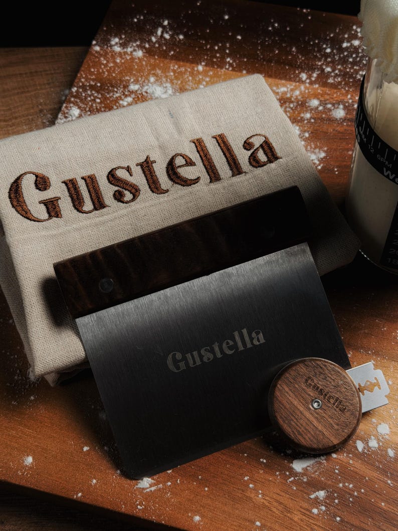 Peut inclure: Un linge en lin beige avec le texte brod&eacute; en marron Gustella, une corne &agrave; p&acirc;tisserie en m&eacute;tal noir avec un manche en bois fonc&eacute; portant le texte grav&eacute; Gustella, et une lame &agrave; pain ronde en bois fonc&eacute; avec une lame en m&eacute;tal et le texte Gustella.