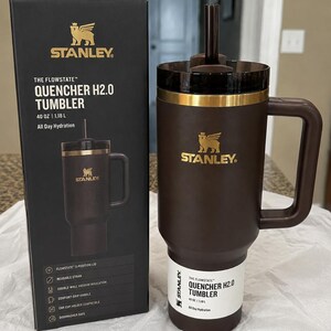 K&ouml;nnte beinhalten: Ein brauner Stanley Quencher H2.0 Becher mit Griff und Strohhalm, neben seiner schwarzen Box. Der Becher hat ein goldenes Band und das Stanley-Logo. Die Box zeigt den Becher und Produktmerkmale.
