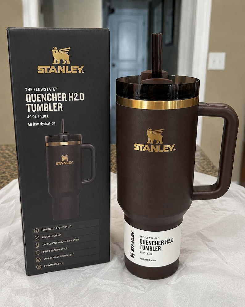 K&ouml;nnte beinhalten: Ein brauner Stanley Quencher H2.0 Becher mit Griff und Strohhalm, neben seiner schwarzen Box. Der Becher hat ein goldenes Band und das Stanley-Logo. Die Box zeigt den Becher und Produktmerkmale.