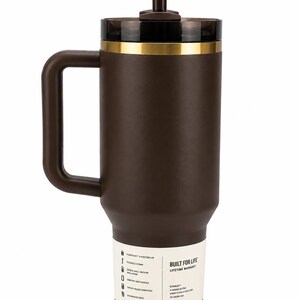 K&ouml;nnte beinhalten: Ein brauner isolierter Becher mit Griff und Strohhalm. Der Becher hat einen schwarzen Deckel mit einem goldfarbenen Band. Ein wei&szlig;es Etikett an der Seite lautet "Built for Life" und listet Funktionen auf. Der Becher ist f&uuml;r Getr&auml;nkehalter im Auto geeignet.