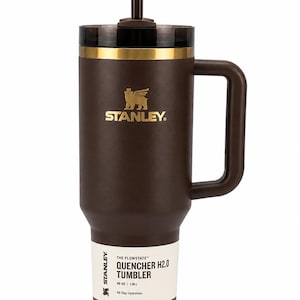K&ouml;nnte beinhalten: Ein brauner Stanley Quencher H2.0 Becher mit Griff und Strohhalm. Der Becher hat einen goldenen Rand und das Stanley-Logo in Gold. Das Etikett lautet "40 oz | 1,18 L" und "All Day Hydration."