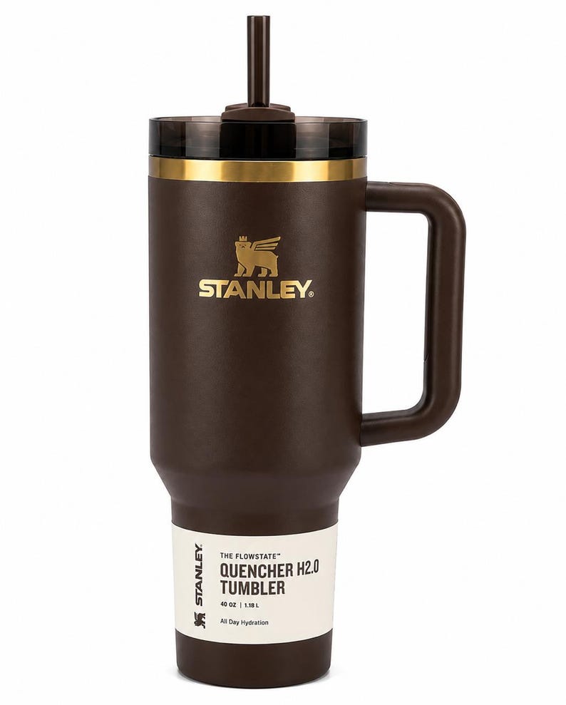 K&ouml;nnte beinhalten: Ein brauner Stanley Quencher H2.0 Becher mit Griff und Strohhalm. Der Becher hat einen goldenen Rand und das Stanley-Logo in Gold. Das Etikett lautet "40 oz | 1,18 L" und "All Day Hydration."