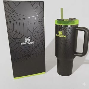 May include: Black Stanley tumbler with a handle, green accents, and a straw, next to a matching box. Both the tumbler and box feature a spiderweb design and the Stanley logo. The tumbler is designed for cold beverages.