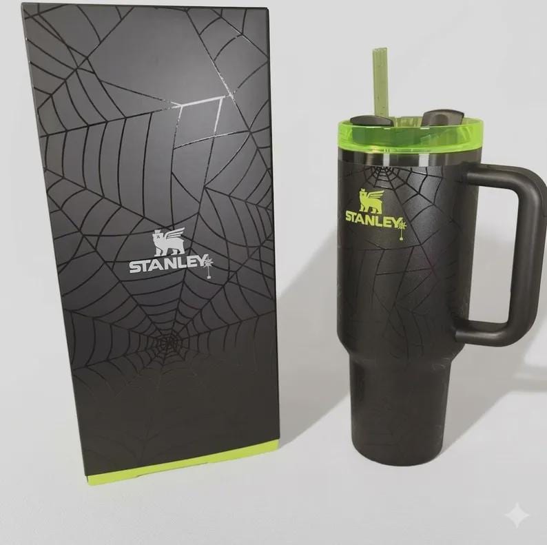 May include: Black Stanley tumbler with a handle, green accents, and a straw, next to a matching box. Both the tumbler and box feature a spiderweb design and the Stanley logo. The tumbler is designed for cold beverages.