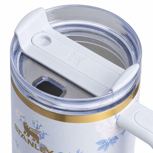 May include: Close-up of a white and floral Stanley travel mug. The mug features a white lid with a sliding closure, a gold band, and a handle. The mug's body is white with a floral pattern in blue and white.