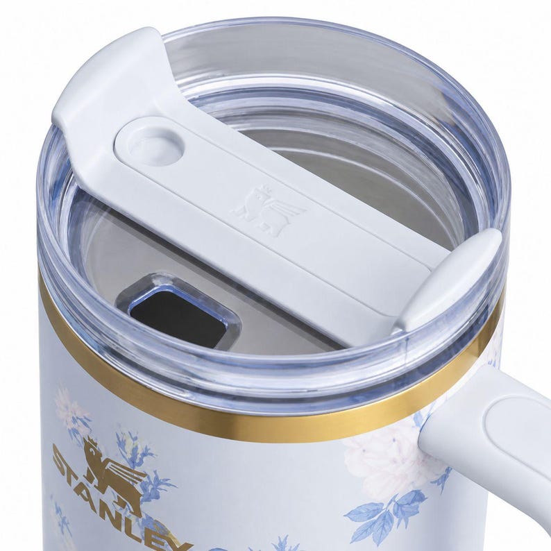 May include: Close-up of a white and floral Stanley travel mug. The mug features a white lid with a sliding closure, a gold band, and a handle. The mug's body is white with a floral pattern in blue and white.