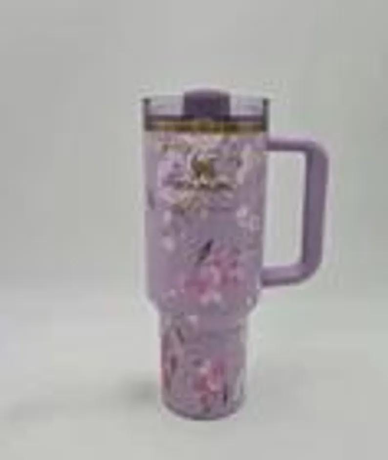 May include: A lavender insulated tumbler with a handle and a floral design. The tumbler has a clear lid and a matching lavender handle. The floral pattern includes pink and white flowers.