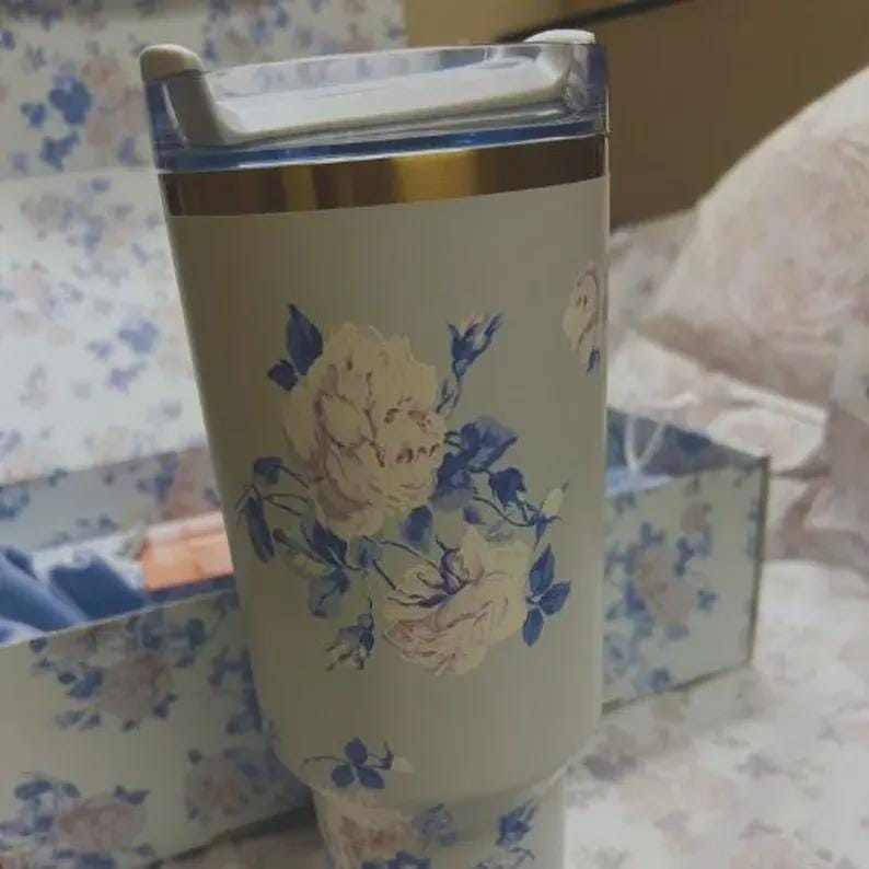 Puede incluir: Un vaso verde claro con un dise&ntilde;o floral con rosas color crema y hojas azules. El vaso tiene una banda dorada cerca de la parte superior y una tapa transparente. El dise&ntilde;o se repite en una caja a juego en el fondo.
