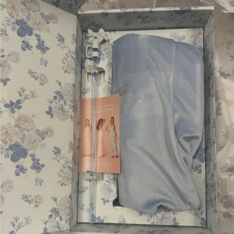 Puede incluir: Una caja abierta con un dise&ntilde;o floral en blanco y azul, que contiene una prenda azul claro, un elemento decorativo largo y transparente y un folleto. El folleto presenta a tres mujeres con vestidos. El contenido de la caja es visible.