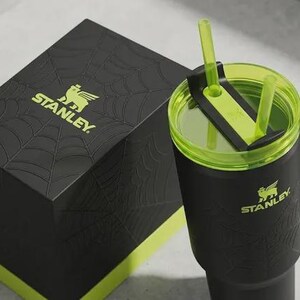 May include: Black Stanley tumbler with a green lid and straw, next to a matching black box. The box and tumbler feature the Stanley logo in green. The box has a spiderweb design. The tumbler has a handle.