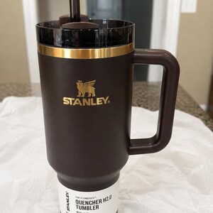 K&ouml;nnte beinhalten: Ein brauner Stanley Quencher H2.0 Becher mit Griff und Strohhalm. Der Becher hat ein goldenes Band am oberen Rand und das Stanley-Logo in Gold. Das Etikett lautet "Quencher H2.0 Tumbler" und "All Day Hydration". Fassungsverm&ouml;gen 1,18 l.