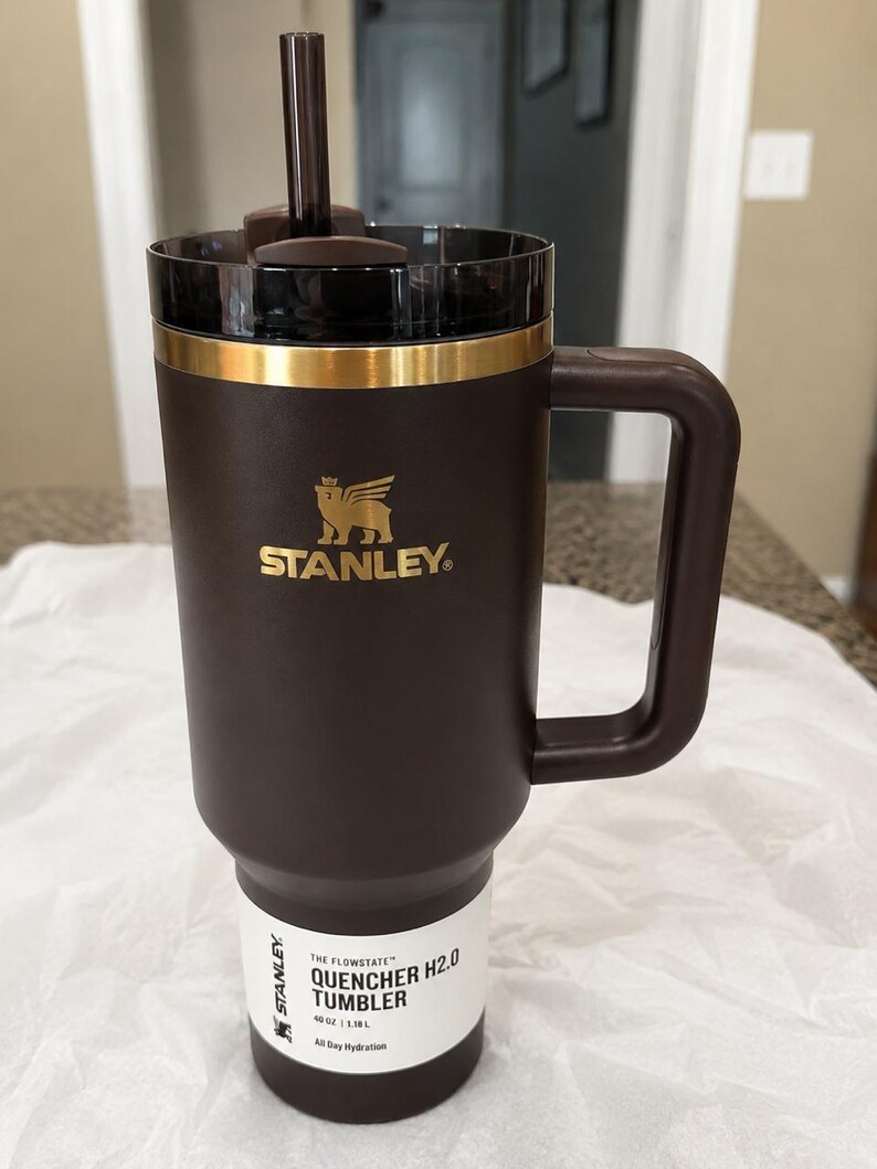 K&ouml;nnte beinhalten: Ein brauner Stanley Quencher H2.0 Becher mit Griff und Strohhalm. Der Becher hat ein goldenes Band am oberen Rand und das Stanley-Logo in Gold. Das Etikett lautet "Quencher H2.0 Tumbler" und "All Day Hydration". Fassungsverm&ouml;gen 1,18 l.
