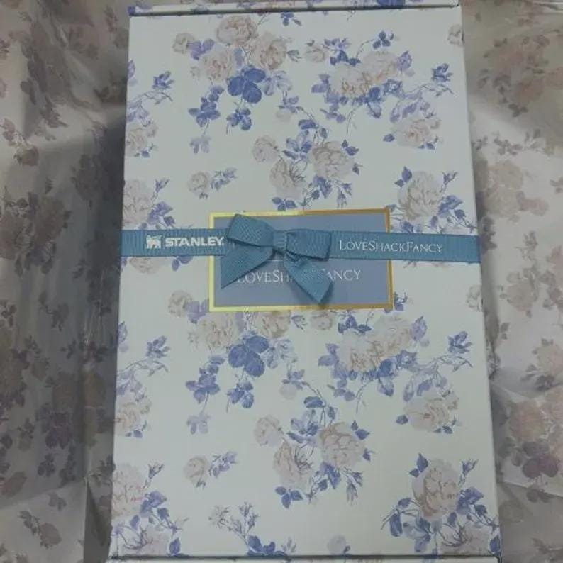 Puede incluir: Una caja de regalo rectangular con un estampado floral en blanco, azul y beige. Una cinta azul con un lazo envuelve la caja, con el texto "STANLEY" y "LOVESHACKFANCY". La caja es probablemente para una ocasi&oacute;n especial.