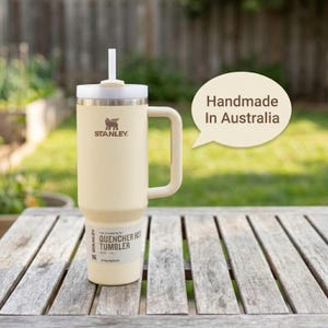 Pu&ograve; includere: Bicchiere Stanley Quencher H2.0 Flowstate color crema con manico e cannuccia. Il bicchiere ha il logo Stanley e la scritta "Handmade In Australia". Il bicchiere &egrave; su un tavolo di legno.