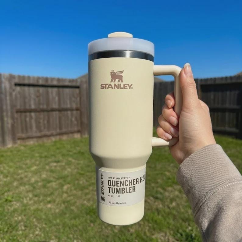Pu&ograve; includere: Un bicchiere Stanley Quencher H2.0 Flowstate color crema con manico, su uno sfondo di erba verde e una staccionata in legno. Il bicchiere ha un coperchio grigio chiaro e il logo Stanley. Il testo sul bicchiere recita "QUENCHER H2.0 TUMBLER".