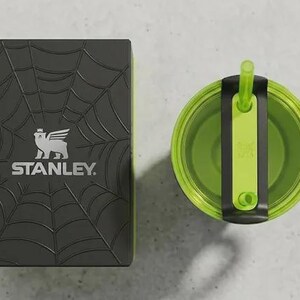 May include: A black Stanley box with a spiderweb design and the Stanley logo next to a green and black tumbler with a straw. The tumbler has a handle and a lid with a straw holder. The items are on a light gray surface.