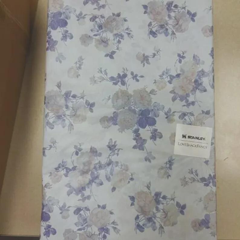 Puede incluir: Tela estampada floral con fondo claro y dise&ntilde;o repetido de rosas y hojas en tonos blancos y morados. La tela tiene una est&eacute;tica suave y rom&aacute;ntica. La etiqueta dice "STANLEY LOVESHACKFANCY".