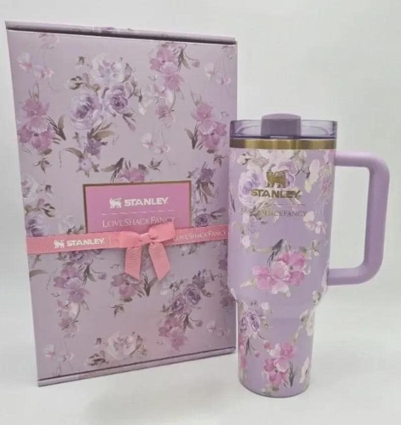 May include: A Stanley Loveshackfancy floral-patterned tumbler and gift box set. The tumbler is lavender with a handle and a lid. The gift box is lavender with a floral design and a pink ribbon. The Stanley logo is visible.