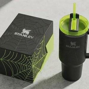May include: Black Stanley tumbler with a green lid and straw, next to a black box with a spiderweb design. The box and tumbler have the Stanley logo. The tumbler has a handle.