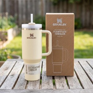 Pu&ograve; includere: Un bicchiere Stanley Quencher H2.0 Flowstate color crema con manico e cannuccia bianchi, accanto alla sua scatola di cartone. Il bicchiere ha il logo Stanley e il testo "40 oz / 1,18L". La scatola mostra anche il logo Stanley e i dettagli del prodotto.
