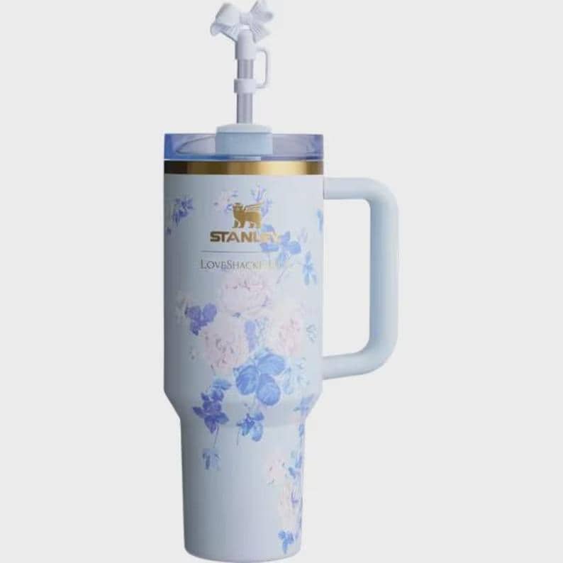 May include: A light blue Stanley Loveshack Fancy tumbler with a handle and a straw. The tumbler is decorated with a floral pattern in shades of blue and white. The top has a clear lid and a white bow on the straw.