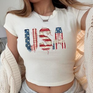 May include: Cream-colored crop top with the word "USA" in a distressed American flag design. The letters are in red, white, and blue, with a frayed edge effect. The top has short sleeves and a round neckline.