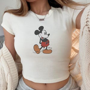 May include: Cream-colored crop top featuring a classic Mickey Mouse graphic. The cartoon character is depicted in black, red, and tan, with the Disney logo below. The top has short sleeves and a rounded neckline.