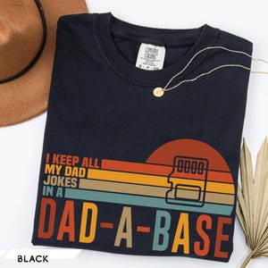 May include: Black t-shirt with a retro sunset design and the text "I KEEP ALL MY DAD JOKES IN A DAD-A-BASE." A gold necklace and a brown hat are also visible. The shirt is a Comfort Colors brand.